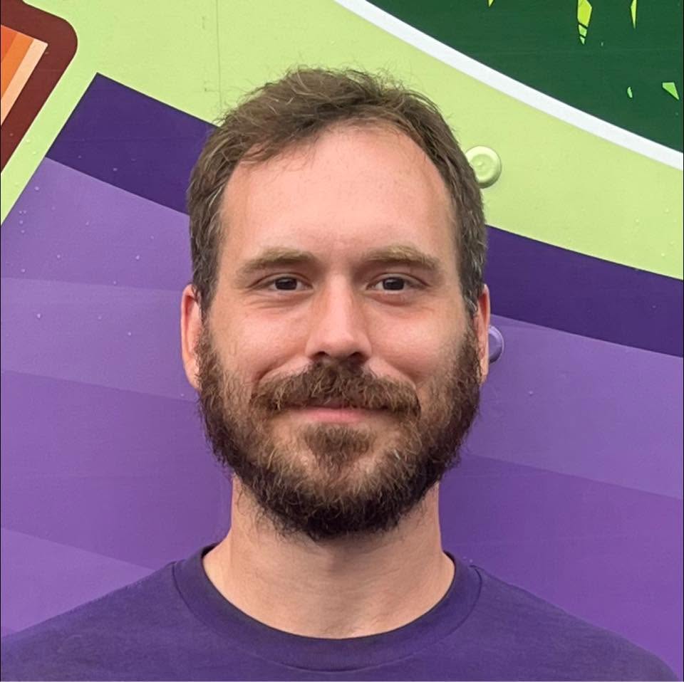 Man with a brown beard and short hair smiles at the camera against a bright, colorful purple-green background.